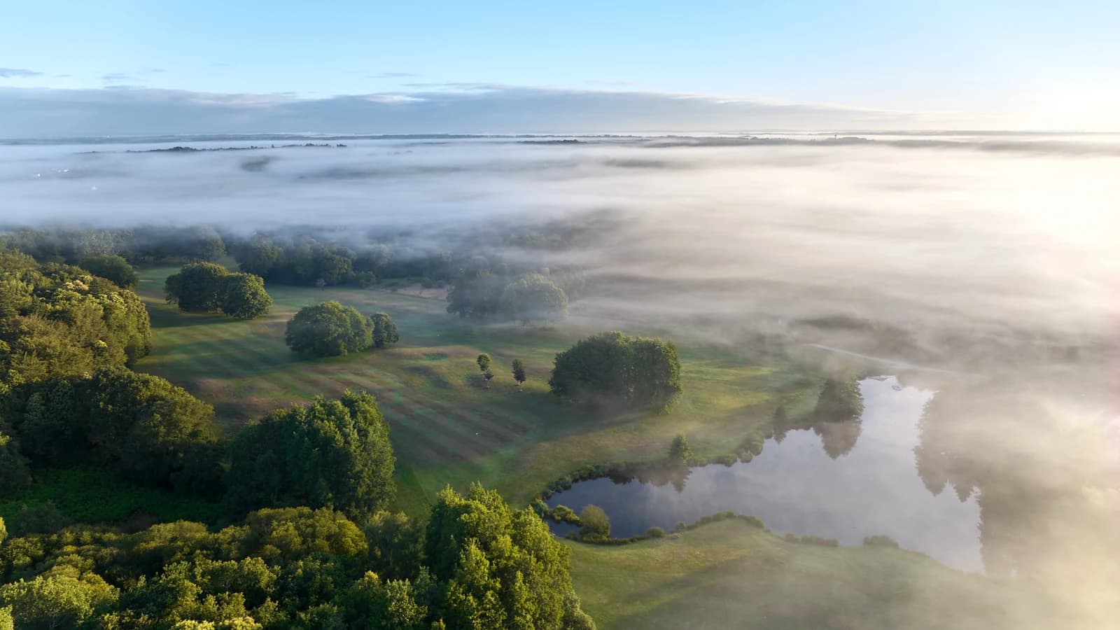 Vue aérienne du parcours boisé et de ses pièces d’eau au Golf Bluegreen Val Quéven, sous une brume matinale.