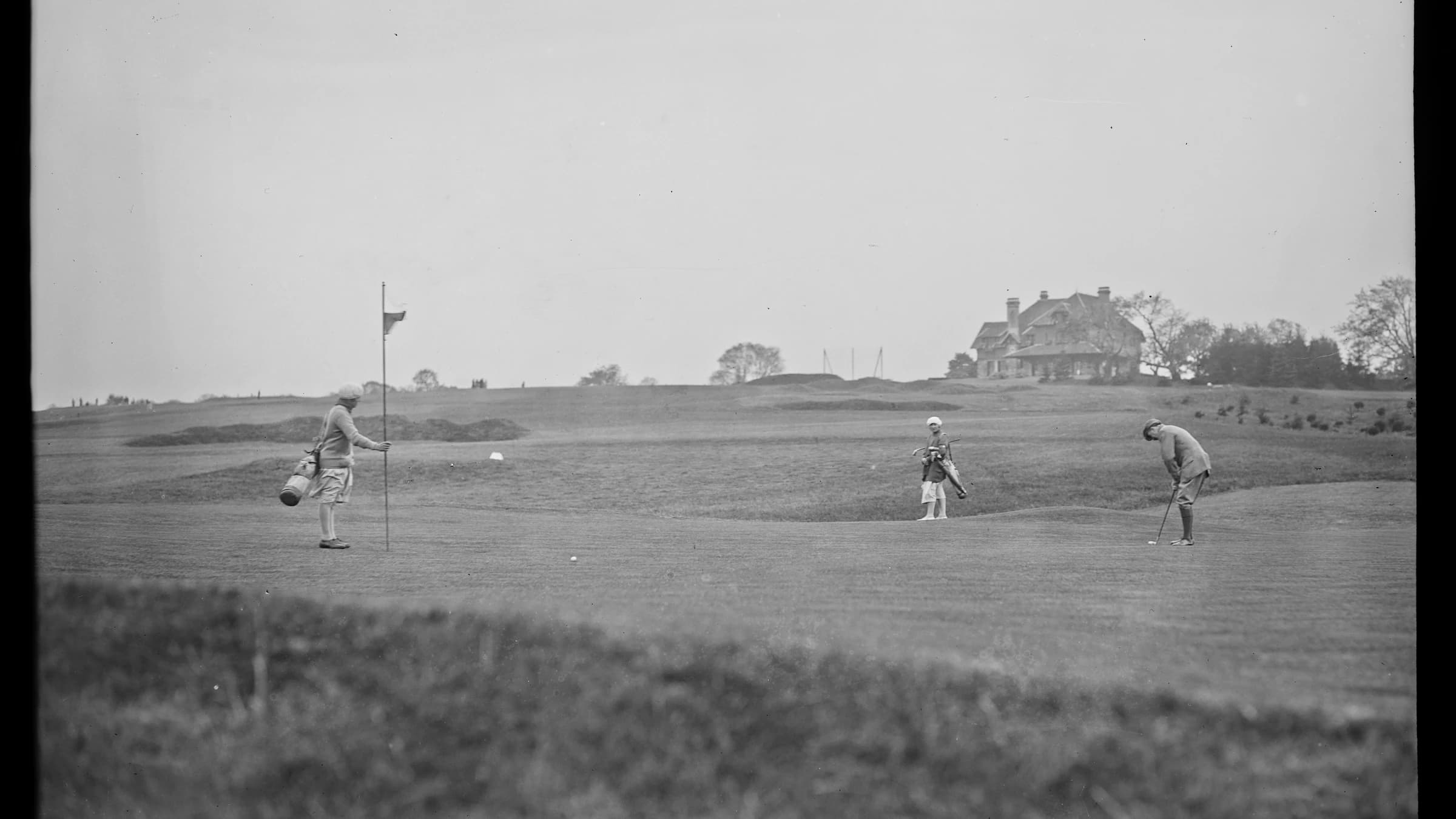 Vue d’archive du green du Golf de La Boulie pendant le championnat de France amateur de 1927, avec le drapeau, trois joueurs et le clubhouse au loin.