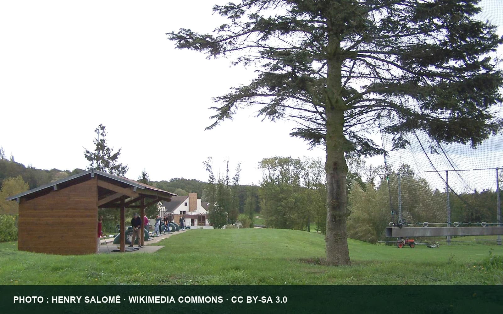 Vue du golf de Feucherolles avec un grand arbre, une zone engazonnée et les bâtiments du club.